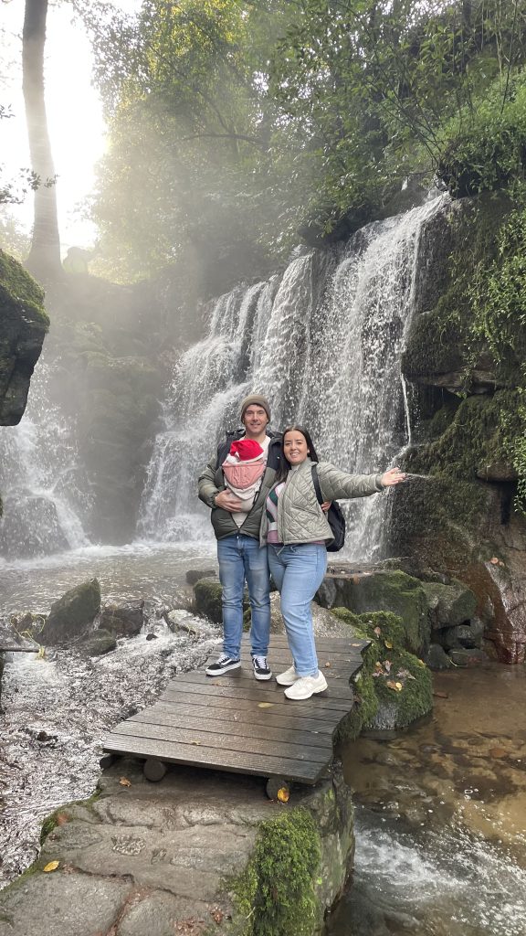 Pareja de padres jóvenes con el bebé en porteo. En la naturaleza a los pies de una cascada.
